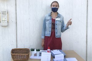 Young white woman in a face mask stands at a check-in table with thumbs up